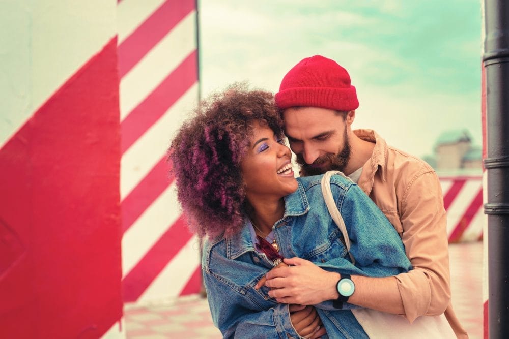 Handsome bearded man kindly smiling and hugging his happy cheerful girlfriend while standing behind her back