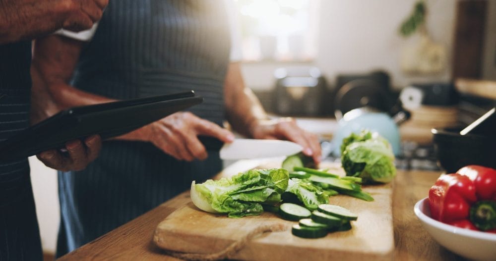 Hands, tablet and salad recipe in kitchen with help, support and teaching at home for healthy food. People cooking lunch on digital tech for nutrition tips with lettuce, cucumber and green vegetables.