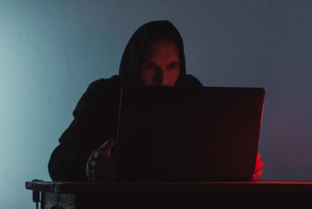 A hooded man committing cyber stalking by looking at stuff on a laptop