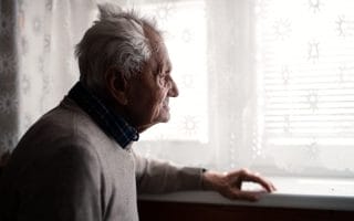 Elderly man standing indoors at home, looking out the window.