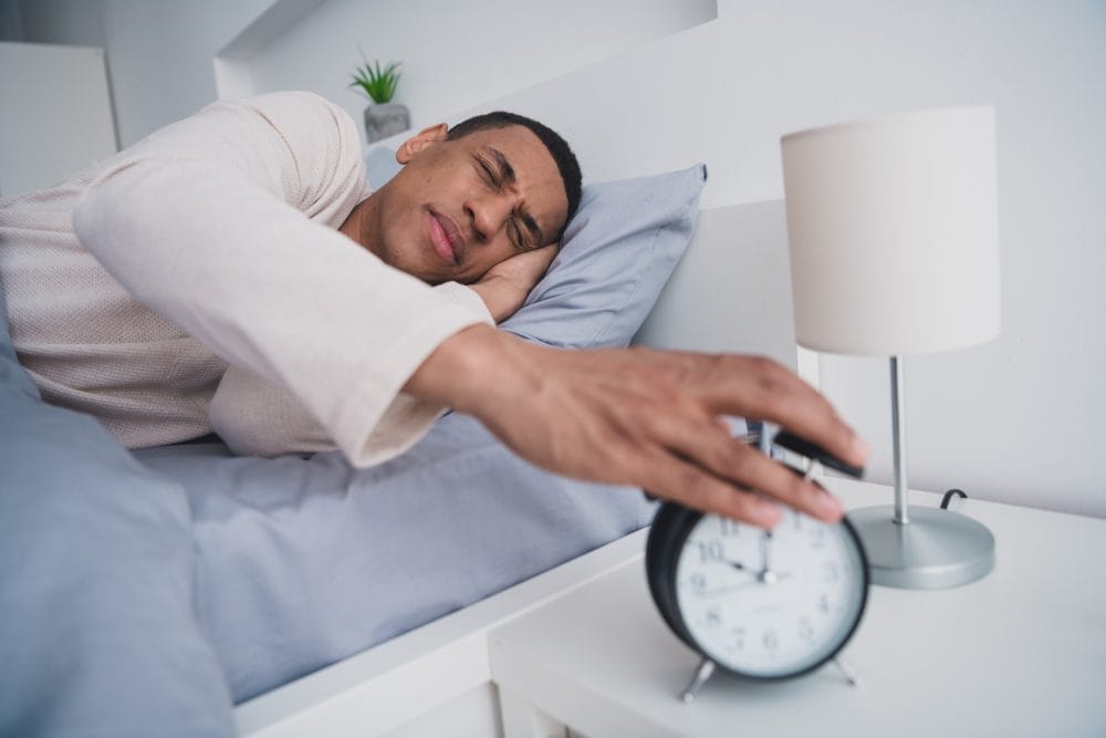 A tired man who didn't get a good night of sleep reaching for his alarm clock
